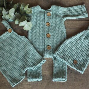 May include: A set of three knitted baby clothes in a light blue color. The set includes a long-sleeved onesie, a pair of overalls, and a hat. All three items have wooden buttons.