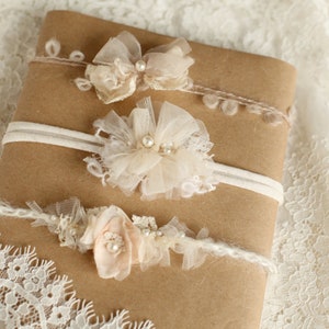 Può includere: Tre fasce per capelli delicate con fiori in tessuto bianco e beige e dettagli di perle. Le fasce sono esposte su uno sfondo di carta marrone.