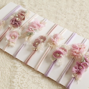 Könnte beinhalten: Eine Auswahl an Baby-Haarbändern mit Stoffblumen in Rosa, Lavendel und Creme. Jedes Haarband ist mit zarter Spitze und Perlen verziert. Die Haarbänder sind auf einer weißen Karte angeordnet, ideal als Geschenk oder für ein Fotoshooting.