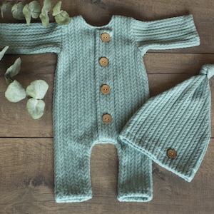 Op de afbeelding: Een lichtblauwe gebreide baby-outfit met een bijpassende muts. De outfit heeft een knoopsluiting en de muts heeft een geknoopte bovenkant.