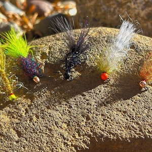 May include: Five different fly fishing lures with colorful feathers and beads. The lures are arranged on a rock surface.