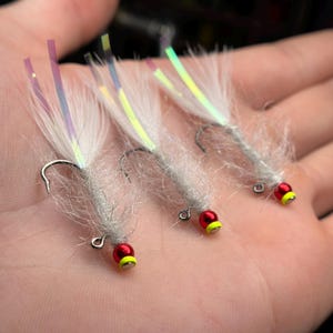 May include: Four white and silver fishing flies with red and yellow beads. The flies have iridescent feathers and are tied on a hook.