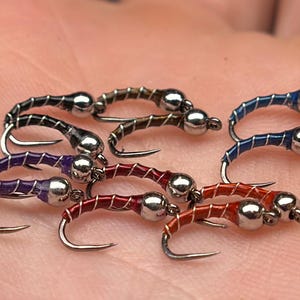 May include: Close-up of several fishing flies in various colors, including black, brown, purple, red, and blue. Each fly has a silver bead head and a curved hook. The flies are designed for fly fishing.