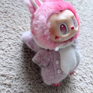 May include: A collectible doll wearing a pink bunny costume with a fluffy hood and ears. The doll has a pale face with red eyes and a mischievous grin. The costume is a soft, knitted material in shades of pink and white.