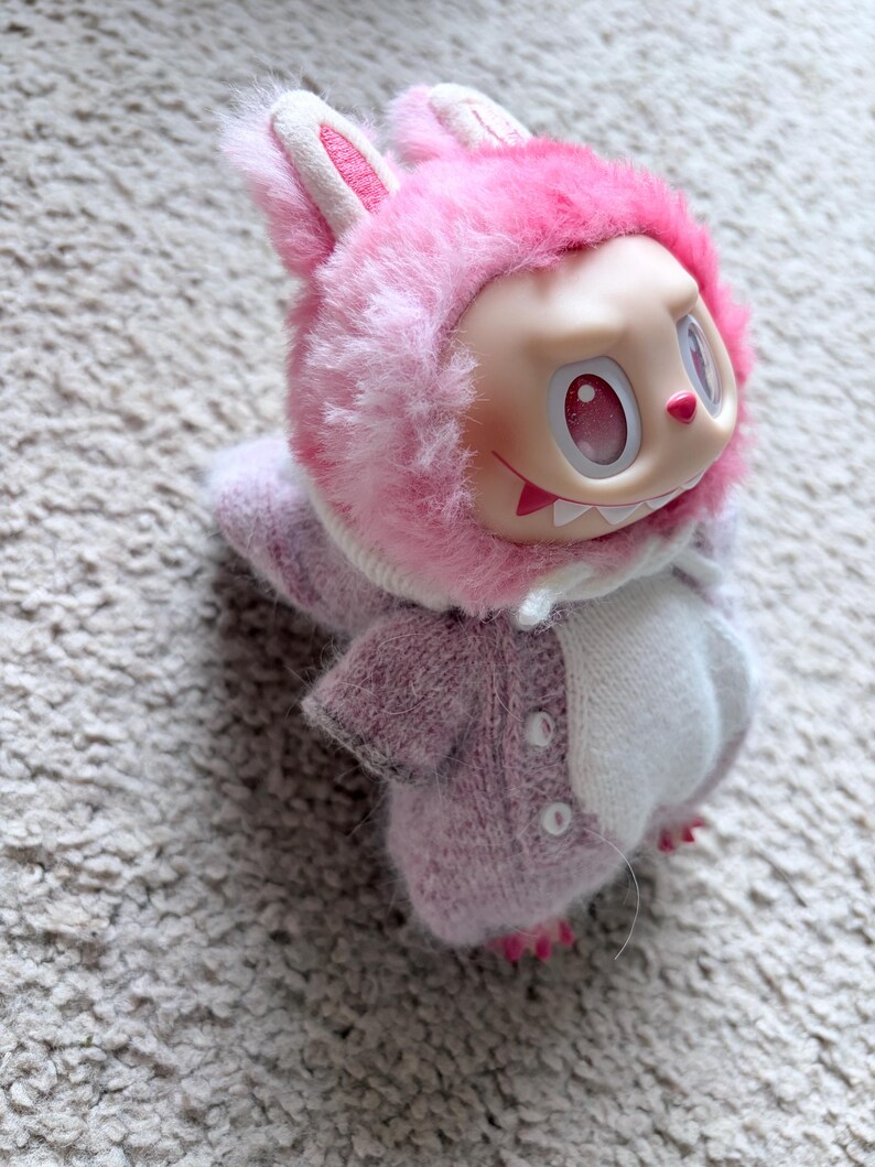 May include: A collectible doll wearing a pink bunny costume with a fluffy hood and ears. The doll has a pale face with red eyes and a mischievous grin. The costume is a soft, knitted material in shades of pink and white.