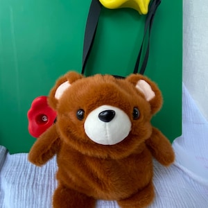 May include: A brown teddy bear-shaped crossbody bag with a white muzzle and black eyes. The bag has a black strap and is set against a green background with a yellow climbing hold and a red flower-shaped hold.