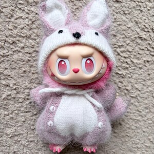May include: A whimsical doll dressed in a pink bunny costume. The doll has a white and pink bunny hat, a pink outfit with white accents, and pink feet. The doll has a mischievous expression with red eyes and a toothy grin.