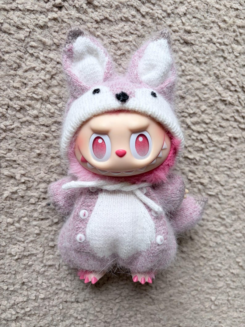 May include: A whimsical doll dressed in a pink bunny costume. The doll has a white and pink bunny hat, a pink outfit with white accents, and pink feet. The doll has a mischievous expression with red eyes and a toothy grin.