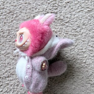 May include: A pink and white plush toy in the shape of a rabbit. The toy has a pink furry hood with white ears, a pink sweater, and a white body. The toy has a smiling face with sharp teeth.