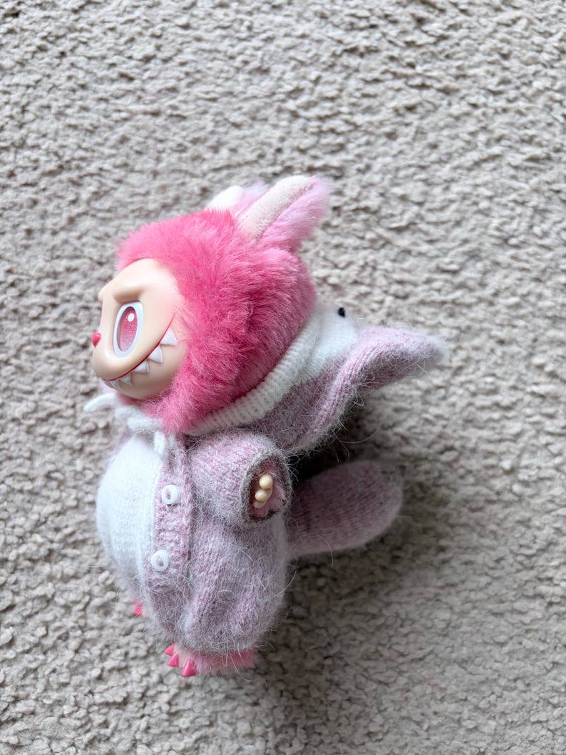 May include: A pink and white plush toy in the shape of a rabbit. The toy has a pink furry hood with white ears, a pink sweater, and a white body. The toy has a smiling face with sharp teeth.