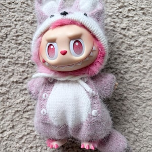 May include: A whimsical doll wearing a pink and white knitted fox hat and matching sweater. The doll has a large head, pink eyes, and a toothy grin. The doll's feet are also pink.