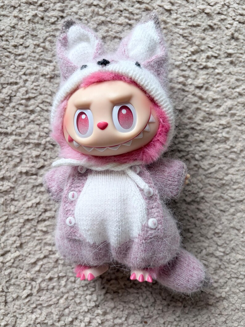 May include: A whimsical doll wearing a pink and white knitted fox hat and matching sweater. The doll has a large head, pink eyes, and a toothy grin. The doll's feet are also pink.