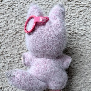 May include: A light purple plush fox toy with a pink keychain clip. The fox has a fluffy texture, gray accents, and a small tail. The keychain clip has the word "POP" printed on it.