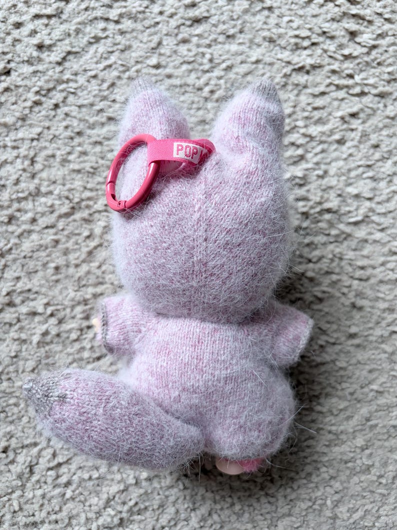 May include: A light purple plush fox toy with a pink keychain clip. The fox has a fluffy texture, gray accents, and a small tail. The keychain clip has the word "POP" printed on it.