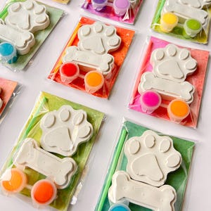 Puede incluir: Un conjunto de adornos de huella de pata y hueso para pintar. Cada conjunto incluye una huella de pata y un hueso de cerámica blanca, dos pequeños botes de pintura, un pincel y una tarjeta colorida. Los botes de pintura son de varios colores, incluyendo naranja, amarillo, azul, verde, rosa y morado.