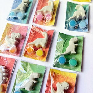 Puede incluir: Pequeñas figuras de dinosaurios blancos empaquetadas con botes de pintura de varios colores. Cada kit incluye un dinosaurio de plástico y un juego de botes de pintura en una bolsa de plástico transparente. El fondo presenta papel estampado colorido.