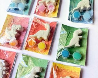 Może przedstawiać: Małe, białe figurki dinozaurów zapakowane z pojemnikami z farbą w różnych kolorach. Każdy zestaw zawiera plastikowego dinozaura i zestaw pojemników z farbą w przezroczystej plastikowej torbie. Tło przedstawia kolorowy papier w wzory.