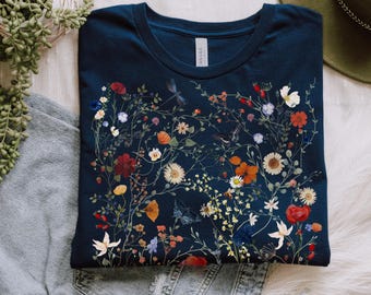 May include: A navy blue t-shirt with a floral design featuring various colorful flowers and greenery. The text "FRENCH NAVY" is printed at the bottom of the shirt. The shirt is laid on top of a pair of jeans.
