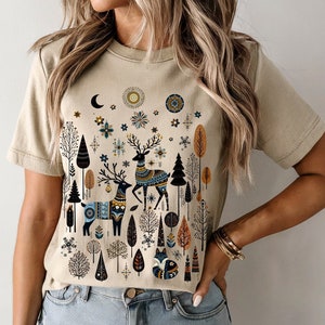 May include: A soft cream-colored t-shirt featuring a whimsical forest scene with deer, trees, and celestial elements. The design incorporates blue, orange, and black accents. The text "SOFT CREAM" is at the bottom right.