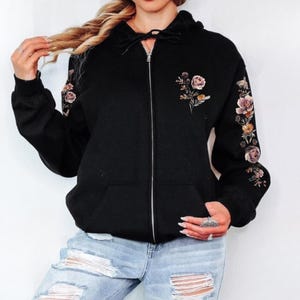 Puede incluir: Sudadera negra con cremallera y bordado floral en el pecho y la manga izquierda. La sudadera con capucha tiene una capucha con cordón y dos bolsillos delanteros. El diseño floral presenta flores rosas, amarillas y naranjas con hojas verdes.