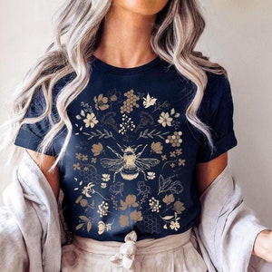 Pode incluir: Camiseta azul marinho com uma ilustração dourada de abelha rodeada por desenhos florais e de favos de mel. A palavra "NAVY" está impressa no canto inferior esquerdo. Uma camiseta gráfica elegante e inspirada na natureza.