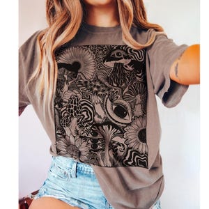 May include: A charcoal gray short-sleeve t-shirt featuring a large, square black graphic print on the front. The design consists of intricate line art illustrations of various mushrooms, sunflowers, and human eyes, all integrated with swirling patterns.