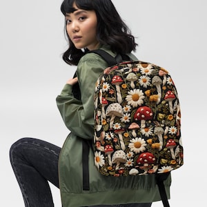 Puede incluir: Una mochila negra con un patrón repetitivo de varios hongos y margaritas blancas. La mochila tiene correas negras y un compartimento principal con cremallera, mostrando un estampado botánico detallado con sombreros de hongos rojos, blancos y bronceados.
