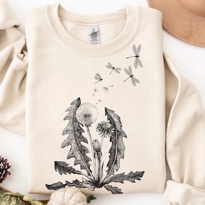 May include: A sand-colored crewneck sweatshirt featuring a black ink-style illustration of a dandelion plant with seeds blowing away and transforming into several dragonflies flying upward toward the neckline of the garment.