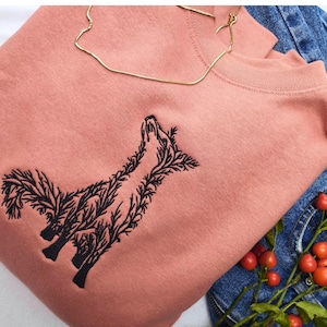 May include: A dusty rose crewneck sweatshirt featuring a black embroidered graphic of a fox composed of intricate tree branches and foliage. The design is centered on the chest of the garment, which is folded alongside blue denim fabric.