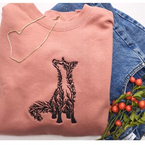 May include: A dusty rose crewneck sweatshirt featuring a black embroidered graphic of a fox composed of intricate branch and leaf patterns. The sweatshirt is paired with blue denim jeans and a small sprig of red berries with green leaves.