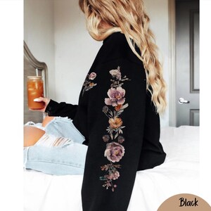 May include: A black long-sleeve sweatshirt featuring a vertical floral print running down the length of the sleeve. The graphic consists of various flowers in shades of muted pink, purple, and orange, arranged in a botanical style on the fabric.