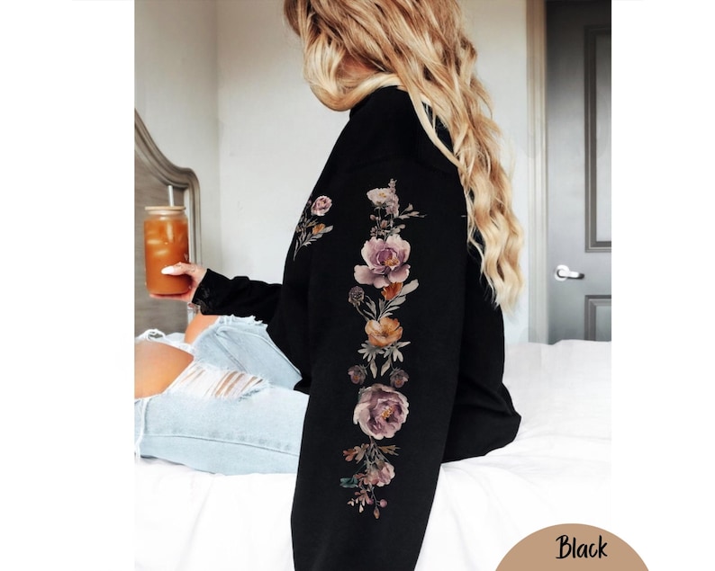 May include: A black long-sleeve sweatshirt featuring a vertical floral print running down the length of the sleeve. The graphic consists of various flowers in shades of muted pink, purple, and orange, arranged in a botanical style on the fabric.