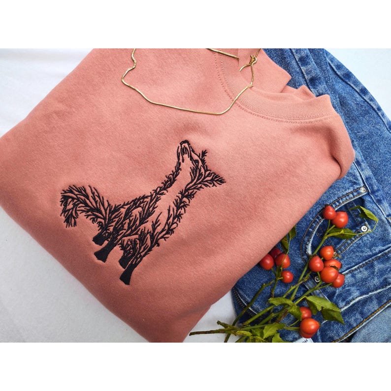 May include: A dusty rose crewneck sweatshirt featuring a black embroidered graphic of a fox composed of intricate tree branches and foliage. The design is centered on the chest of the garment, which is folded alongside blue denim fabric.