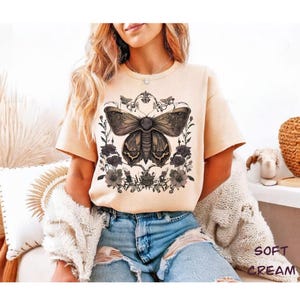 May include: Soft cream-colored t-shirt featuring a detailed illustration of a moth with floral accents. The moth design is in shades of brown and black, surrounded by dark gray flowers and foliage. The shirt has short sleeves and a crew neck.