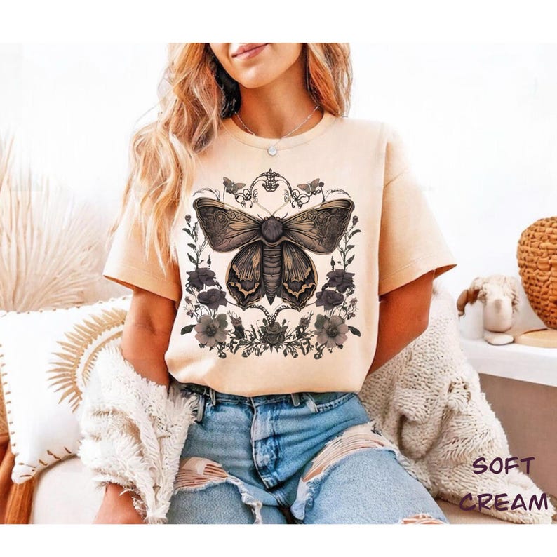 May include: Soft cream-colored t-shirt featuring a detailed illustration of a moth with floral accents. The moth design is in shades of brown and black, surrounded by dark gray flowers and foliage. The shirt has short sleeves and a crew neck.