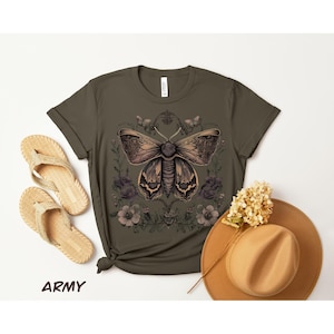 May include: Army green t-shirt featuring a detailed moth illustration with floral accents. The design is in shades of brown and tan. A straw hat and woven sandals are also visible.