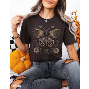 May include: Brown t-shirt featuring a detailed butterfly design with floral accents. The butterfly has intricate wing patterns and is surrounded by dark flowers. The word "BROWN" is printed at the bottom of the image.