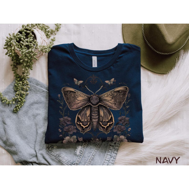 May include: Navy blue t-shirt featuring a detailed illustration of a moth with floral accents. The moth is brown and tan, with intricate wing patterns. The shirt is displayed on a white surface with a green hat and a plant in the background. The word "NAVY" is visible.