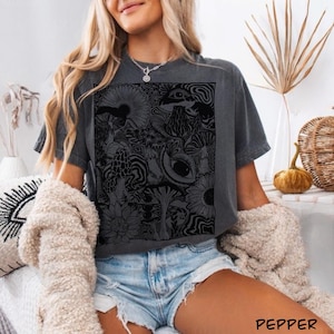 May include: A charcoal gray short-sleeve t-shirt featuring a large, rectangular black graphic print on the front. The design consists of intricate, detailed line art depicting various mushrooms, sunflowers, and a prominent eye surrounded by swirls.