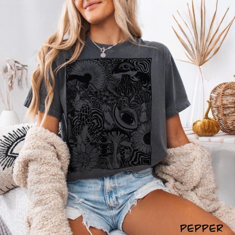 May include: A charcoal gray short-sleeve t-shirt featuring a large, rectangular black graphic print on the front. The design consists of intricate, detailed line art depicting various mushrooms, sunflowers, and a prominent eye surrounded by swirls.