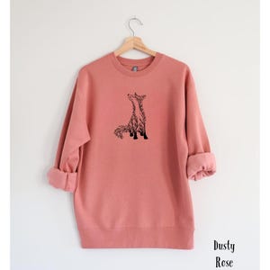 May include: A dusty rose long-sleeve sweatshirt featuring a black line art graphic of a fox on the center chest. The garment has a crew neckline, ribbed cuffs, and a ribbed hem. The text Dusty Rose is printed in black in the bottom right corner.