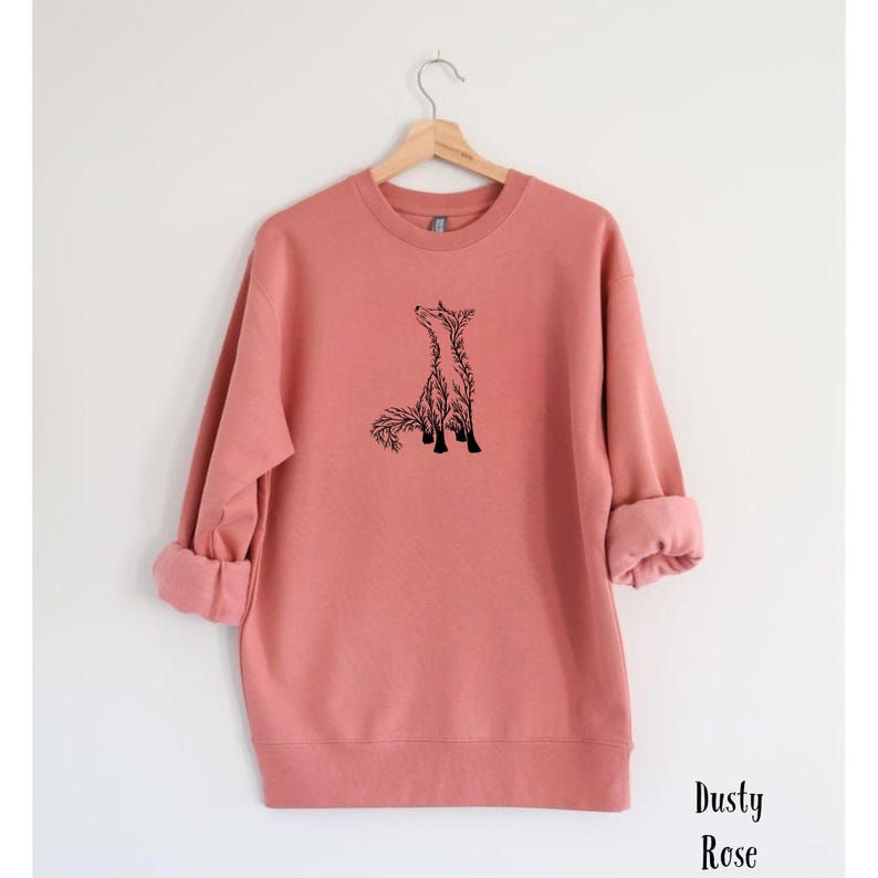 May include: A dusty rose long-sleeve sweatshirt featuring a black line art graphic of a fox on the center chest. The garment has a crew neckline, ribbed cuffs, and a ribbed hem. The text Dusty Rose is printed in black in the bottom right corner.
