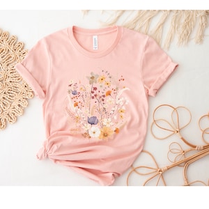May include: A pink t-shirt with a floral print featuring a variety of flowers in shades of yellow, white, blue, and red.