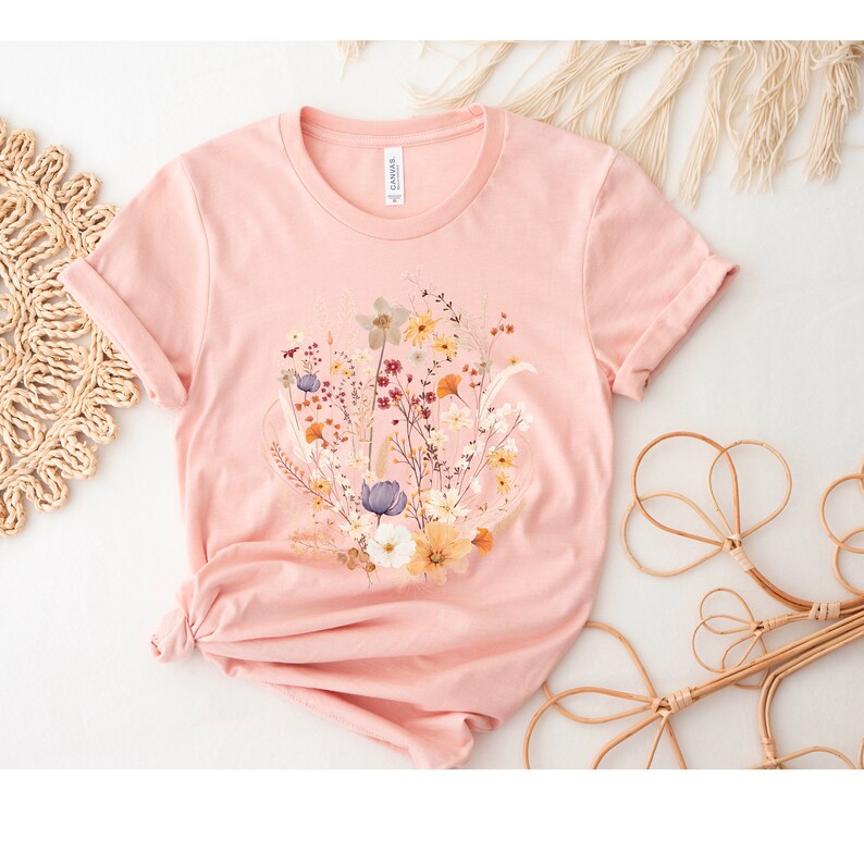 May include: A pink t-shirt with a floral print featuring a variety of flowers in shades of yellow, white, blue, and red.