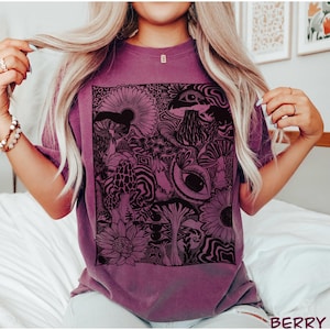 May include: A berry-colored short-sleeve t-shirt featuring a large, rectangular black graphic print on the front. The design consists of intricate illustrations of various mushrooms, sunflowers, and stylized human eyes, all arranged in a dense, detailed pattern.