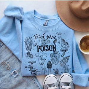 May include: Light blue sweatshirt with the text "Pick your POISON" and various mushroom illustrations in black. The sweatshirt is paired with light-wash denim jeans, a brown hat, and a cup of coffee.