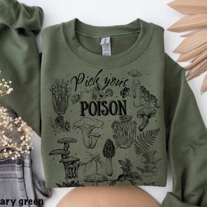 May include: Military green sweatshirt with a black graphic of various mushrooms and the text "Pick your POISON". The sweatshirt is a crew neck style and is made of a soft material. The sweatshirt is folded on a white surface.