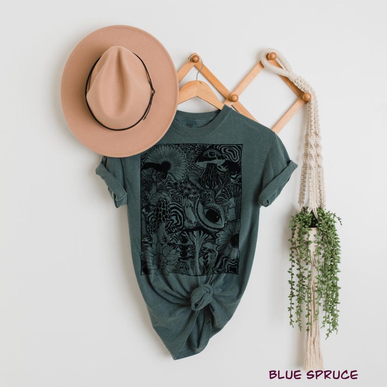 May include: A dark teal short-sleeved t-shirt featuring a black rectangular graphic print of mushrooms, flowers, and an eye. A tan wide-brimmed hat with a black band is positioned above the shirt. The text "BLUE SPRUCE" appears in the bottom right.
