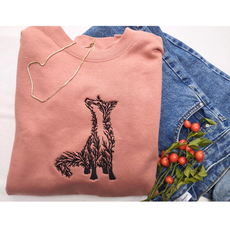May include: A dusty rose crewneck sweatshirt featuring a black embroidered graphic of a fox composed of intricate branch and leaf patterns. The sweatshirt is paired with blue denim jeans and a small sprig of red berries with green leaves.