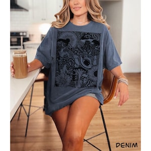 May include: A denim blue short-sleeve t-shirt featuring a large rectangular black graphic print on the front. The design consists of intricate, detailed illustrations of various mushrooms, flowers, swirling patterns, and a prominent eye.